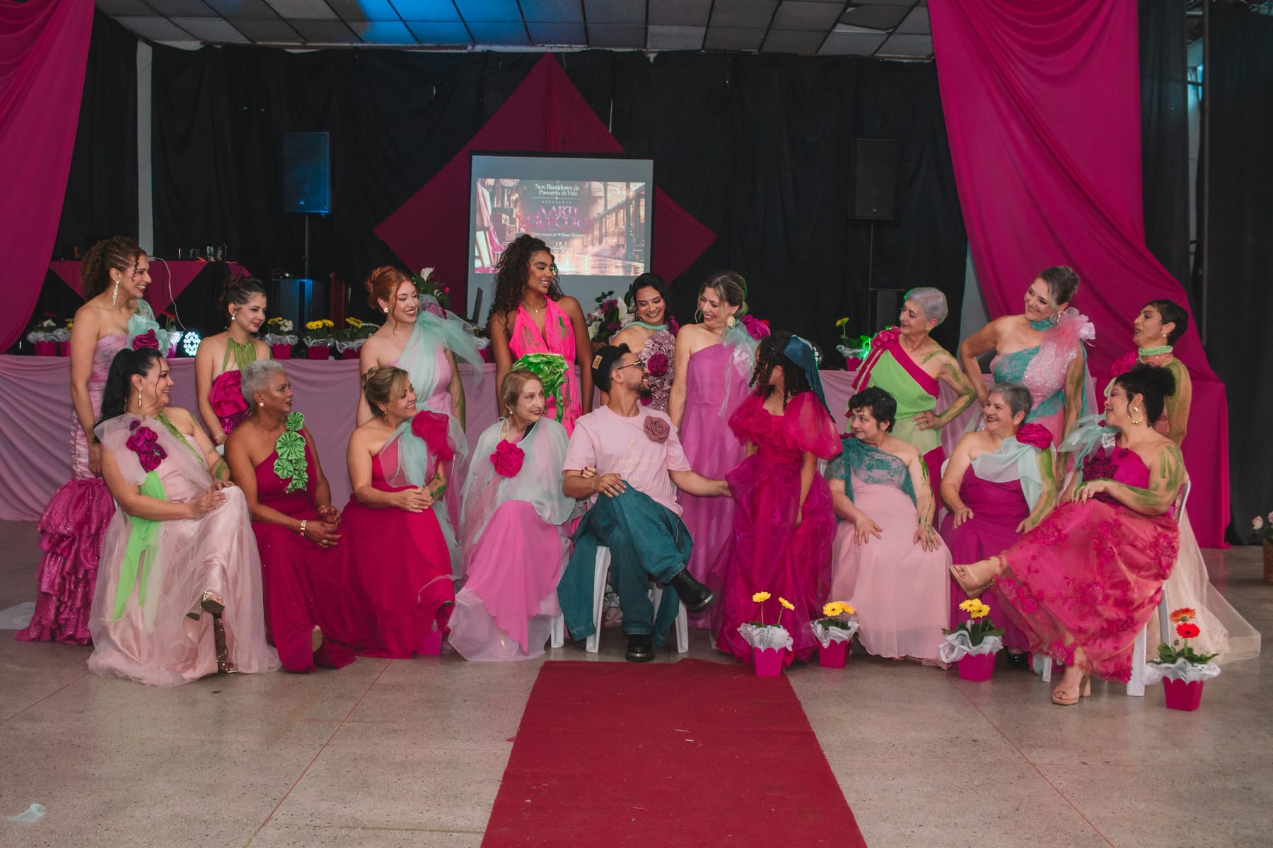 Participantes do desfile sentadas e em pé ao redor do estilista, sorrindo