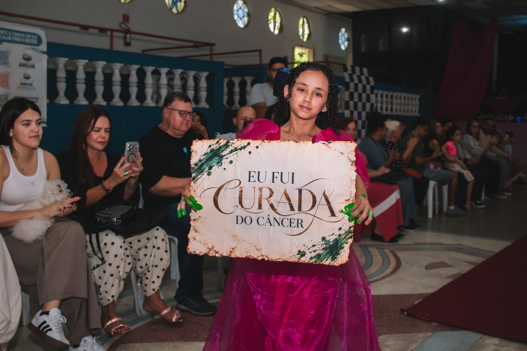Menina de vestido rosa segura uma placa com a frase Eu fui curada do câncer